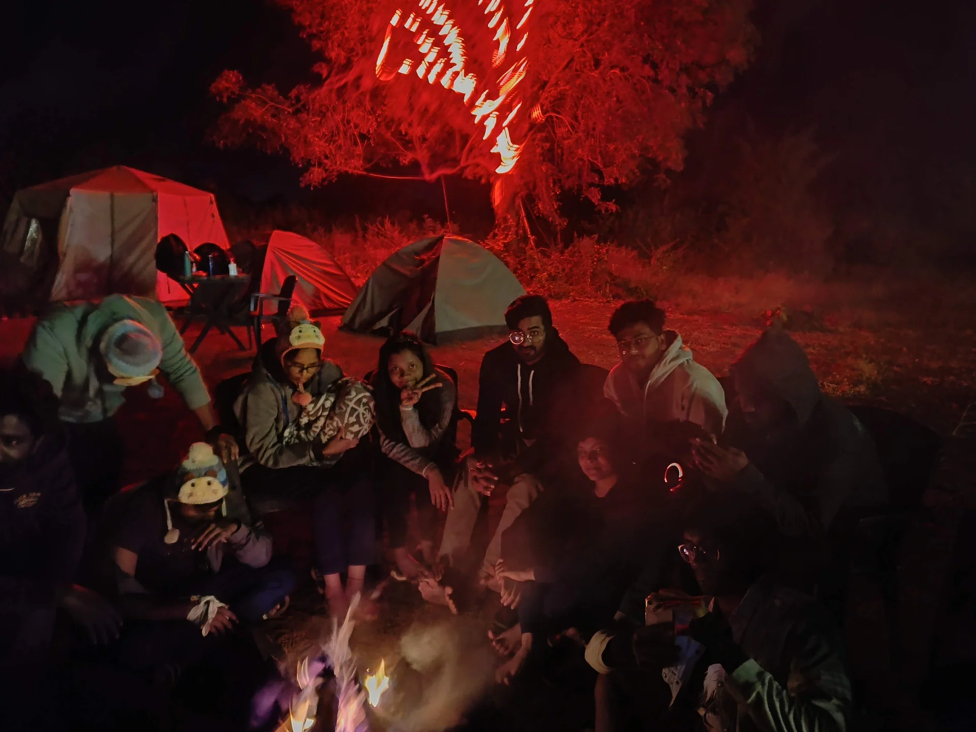 Group around a bonfire with tents and red-lit trees