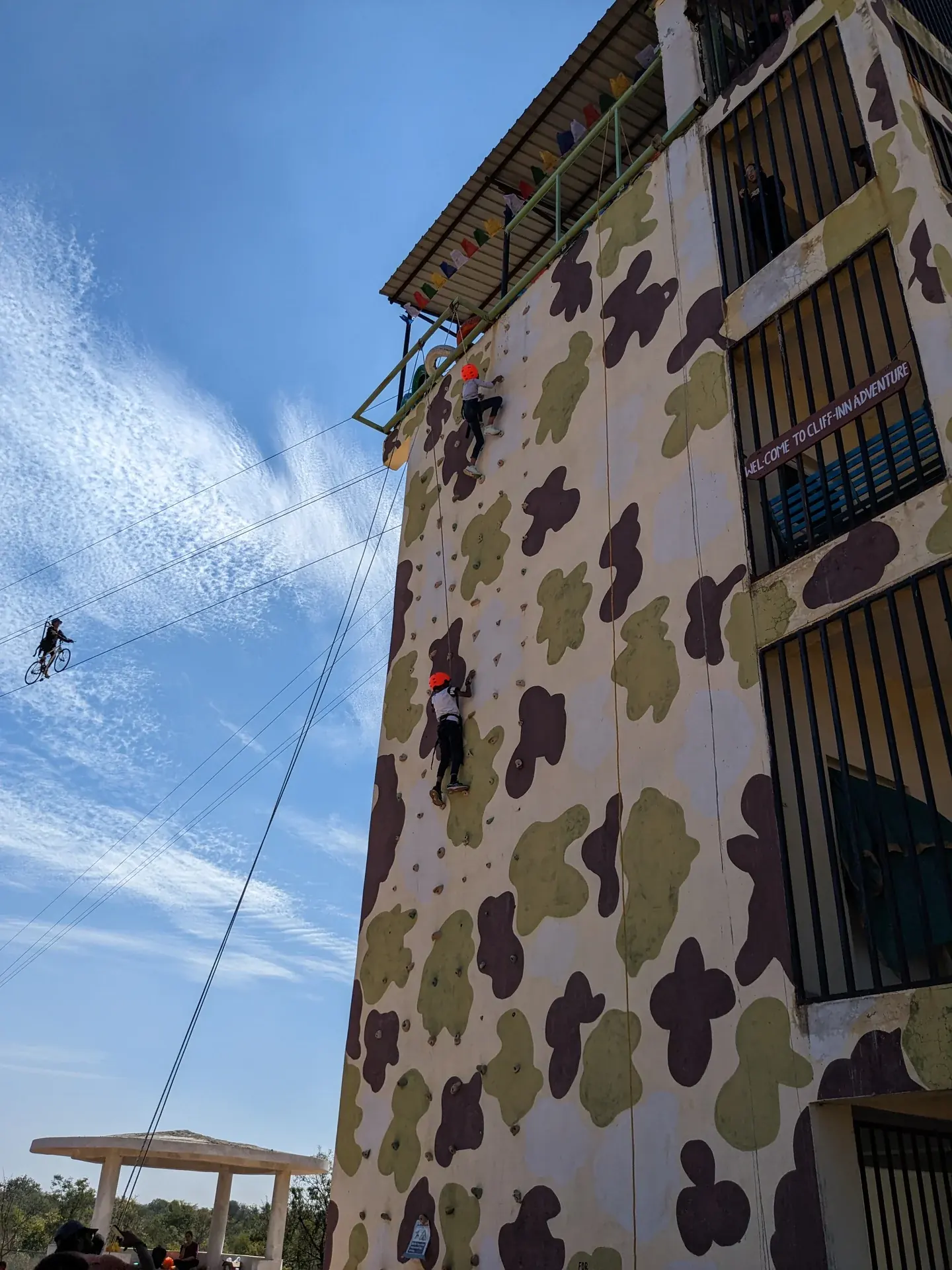 Students scaling a military-pattern rock climbing wall with zipline visible in the background